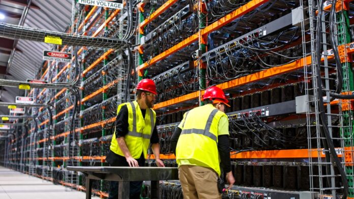 Two technicians wearing PPE walking past racks of equipment in a data center for AI computing in a large, temperature controlled warehouse in a remote location in Stutsman County, North Dakota. One of them is pushing a cart.